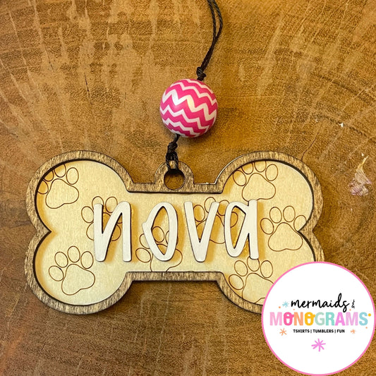 Wooden bone-shaped pet tag with 'nova' engraved, on a wooden background with a pink and white zigzag bead.