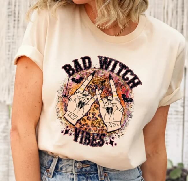 Woman wearing a cream-colored t-shirt with a "Bad Witch Vibes" design featuring tattooed witch hands, leopard print moon, and Halloween symbols like bats and spiderwebs.