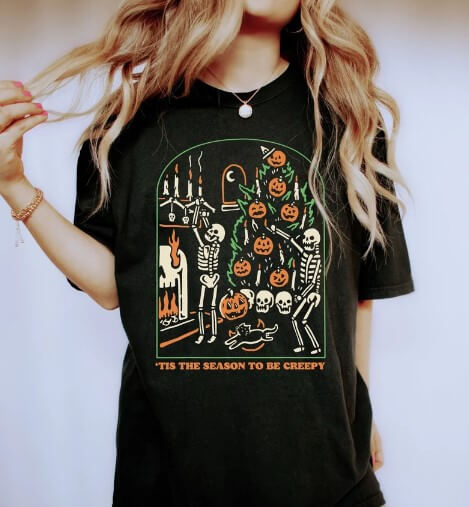 Woman wearing a black Halloween t-shirt with dancing skeletons and a decorated pumpkin tree design, captioned "’Tis the Season to Be Creepy