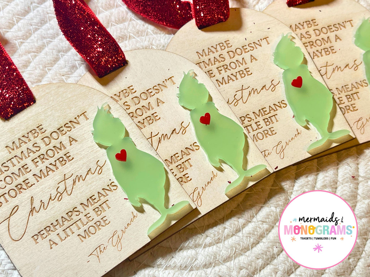 Close-up side angle of multiple handmade Grinch ornaments with red glitter ribbon, engraved holiday quote, and light green acrylic silhouette with a red heart accent.