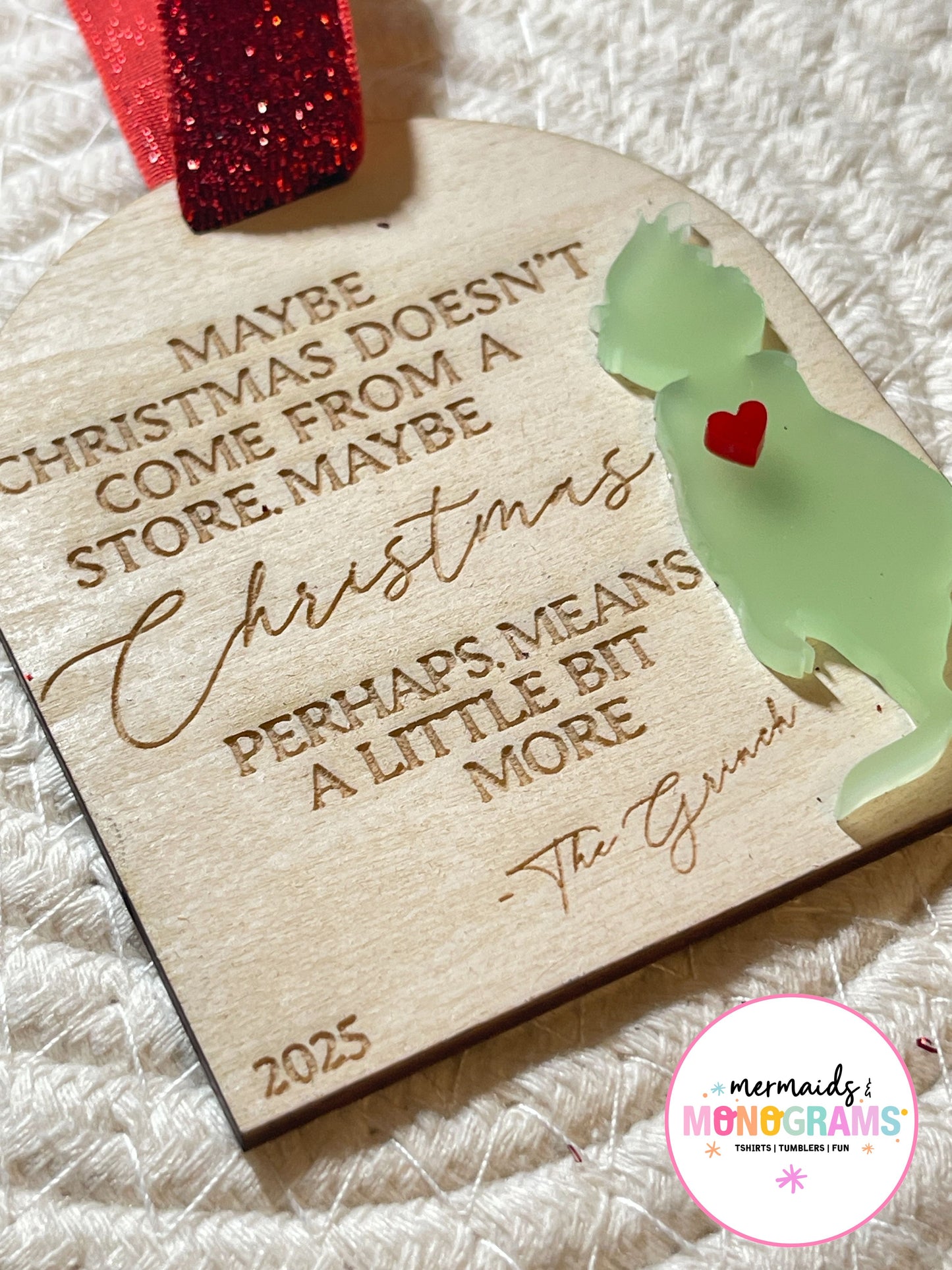 Detailed view of wooden Christmas ornament engraved with Grinch quote and dated 2025, featuring green acrylic figure and sparkling red ribbon.