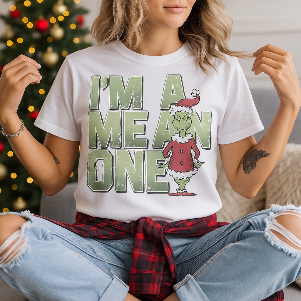 Woman wearing a white t-shirt with large green text that says “I’m A Mean One” next to a cartoon grinch-style character in a red Santa outfit, sitting cross-legged in front of a Christmas tree.