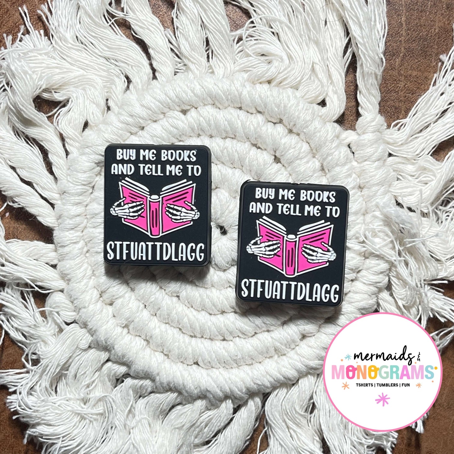 Two black silicone beads with pink designs and text on a white macramé background, featuring the brand 'Mermaids & Monograms'.