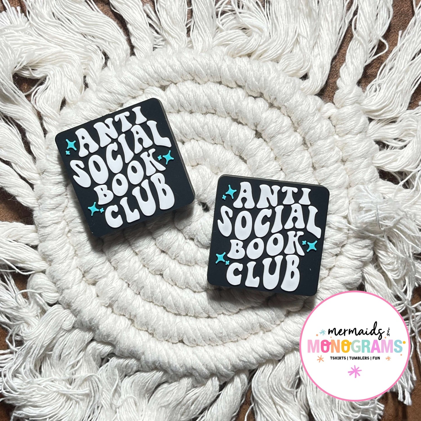 Two black silicone beads with 'Anti Social Book Club' text on a white macramé background.
