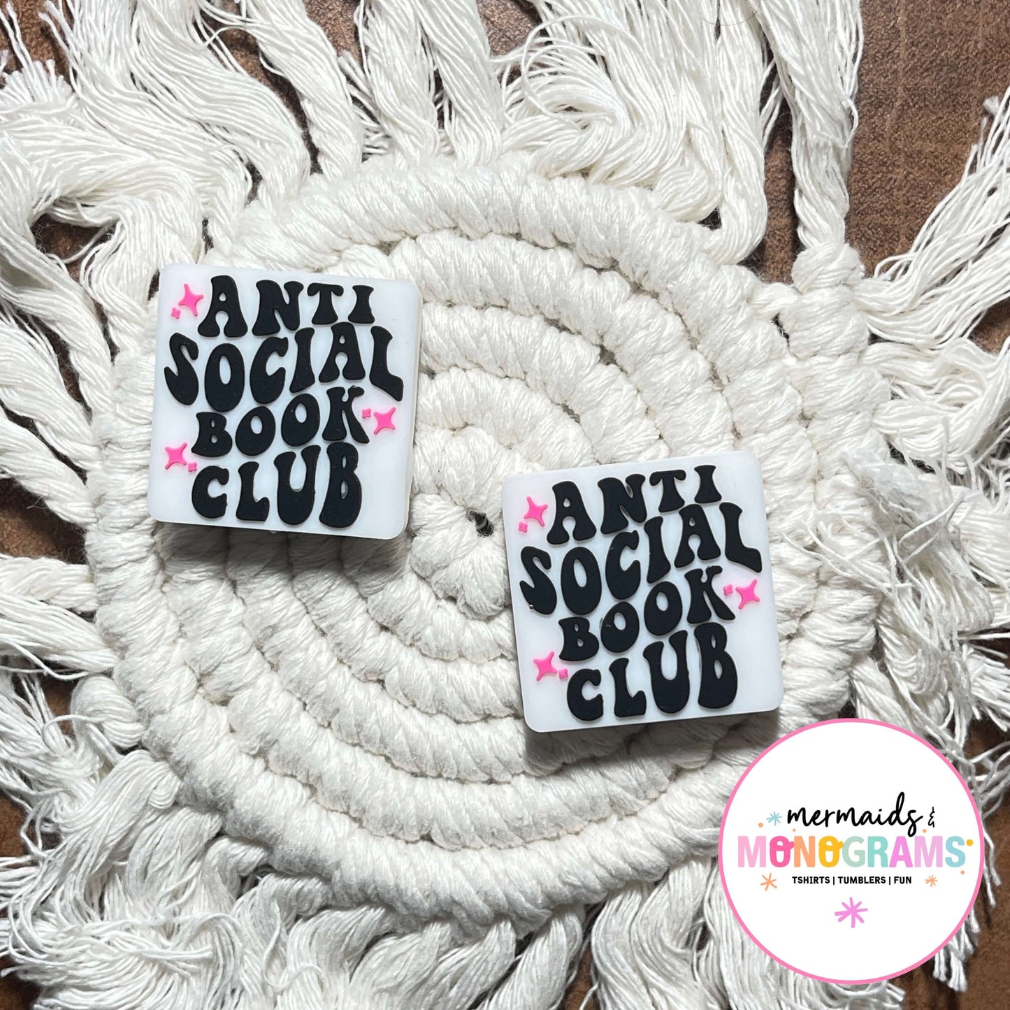 Two 'Anti Social Book Club' beads on a macrame surface with a brand logo in the corner.