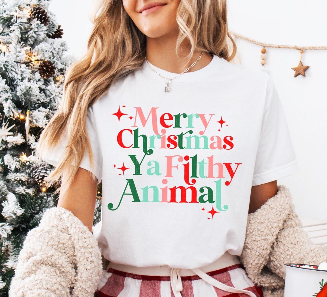 Image of a woman wearing a "Merry Christmas Ya Filthy Animal" T-shirt in retro red, green, and pink fonts, styled with cozy winter accessories and a Christmas tree in the background.