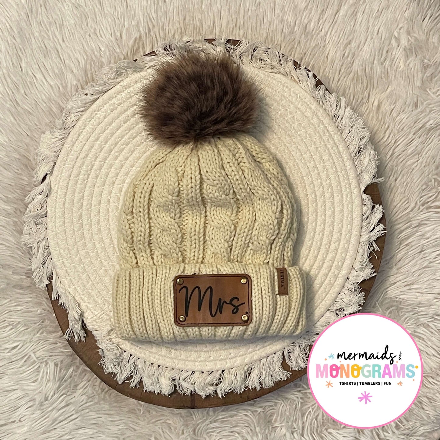 Cream cable knit pom pom beanie with leather "Mrs." patch displayed flat