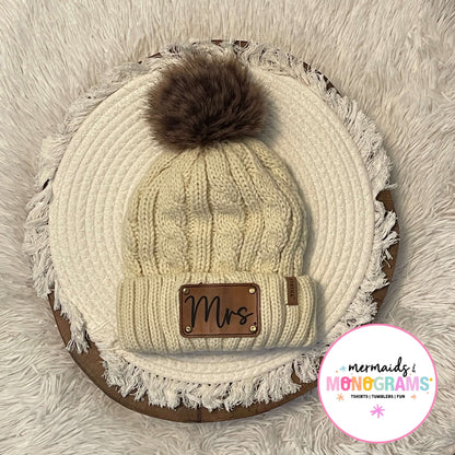 Cream cable knit pom pom beanie with leather "Mrs." patch displayed flat
