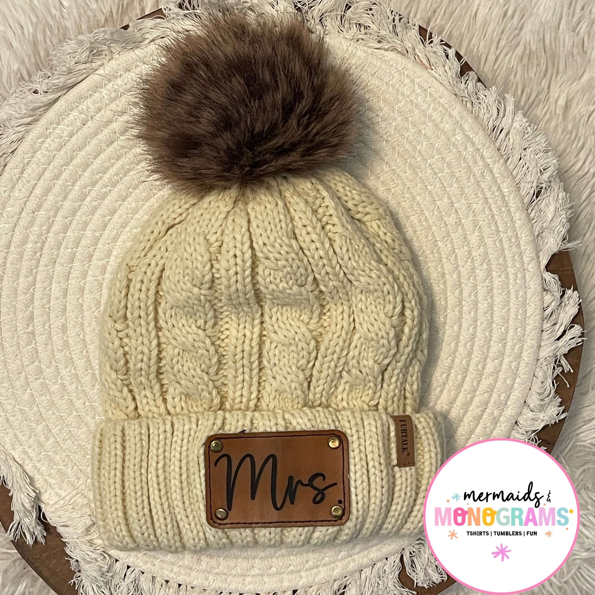 Full view of pom pom hat with neutral faux fur top and brown leather patch