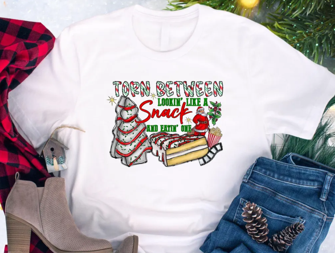 Flat lay of a white Christmas t-shirt with the phrase “Torn Between Lookin’ Like a Snack and Eatin’ One” featuring a Santa, decorated Christmas trees, and snacks, styled with boots, flannel, and pinecones for a festive holiday look.